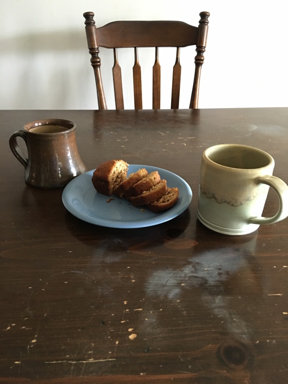 A wooden table set with two coffee/tea mugs and a plate of banana bread slices. A chair along the far side.